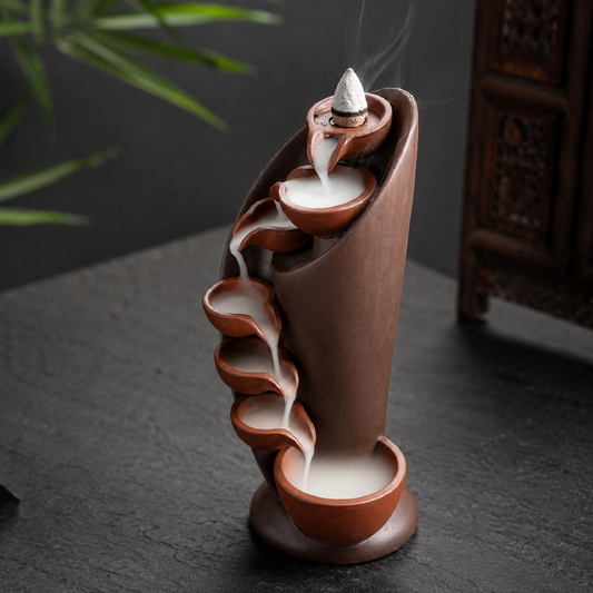 Ceramic waterfall backflow incense burner with cascading smoke placed on a dark tabletop.