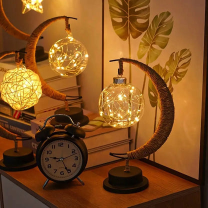 A beautifully designed crescent moon table lamp features a hanging glass orb filled with warm LED fairy lights, creating a soft and inviting glow. Placed on a wooden surface alongside a classic alarm clock, stacked books, and leafy wall decor, the scene highlights a cozy, modern interior aesthetic perfect for bedrooms, study tables, or ambient home décor.