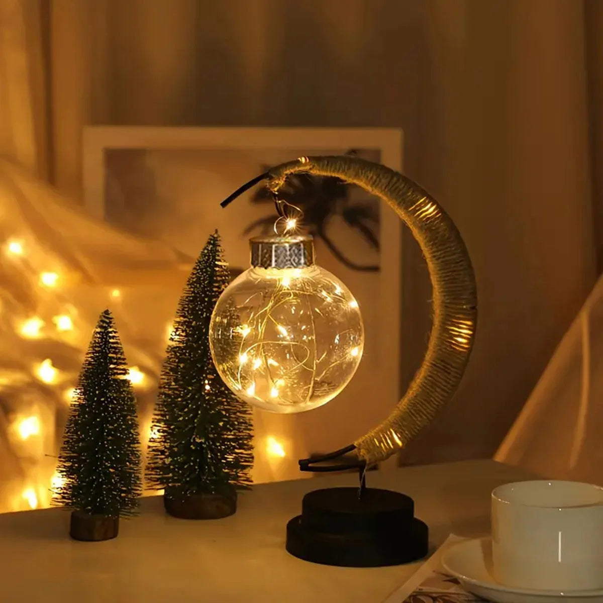 A charming crescent moon table lamp features a hanging glass globe filled with warm fairy lights, creating a soft and inviting glow. Styled with small decorative trees and twinkling background lights, this lamp adds a cozy, festive ambiance to any room—perfect for bedrooms, holiday décor, or relaxing evening settings.