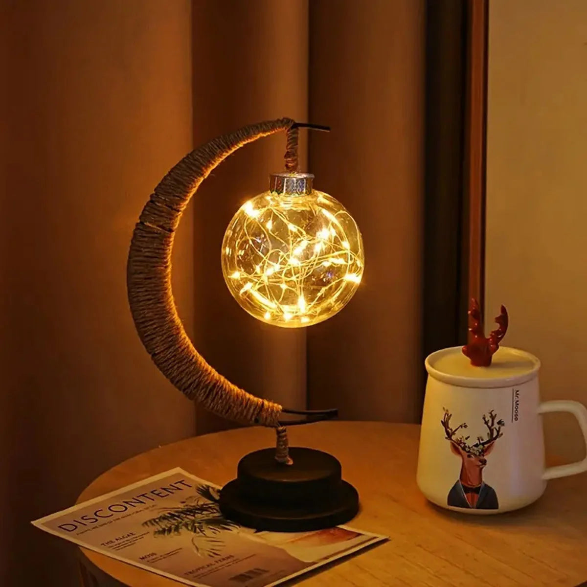 A stylish crescent moon-shaped table lamp features a glowing glass orb filled with warm LED string lights, creating a cozy and ambient atmosphere. Placed on a wooden side table beside a decorative mug and a magazine, with soft curtains in the background, this lamp adds a warm, modern touch to any bedroom or living space.