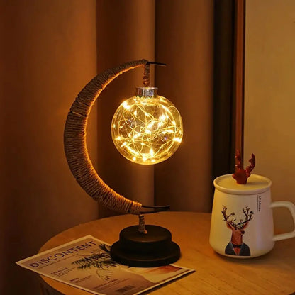 A stylish crescent moon-shaped table lamp features a glowing glass orb filled with warm LED string lights, creating a cozy and ambient atmosphere. Placed on a wooden side table beside a decorative mug and a magazine, with soft curtains in the background, this lamp adds a warm, modern touch to any bedroom or living space.