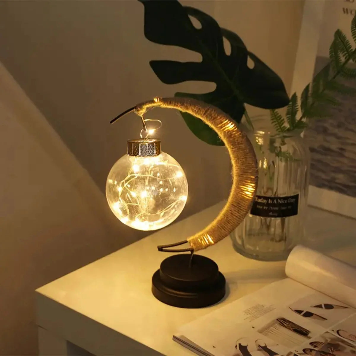 A contemporary crescent moon-shaped table lamp features a hanging glass globe filled with warm glowing fairy lights, creating a soothing and ambient glow. Placed on a minimalist white table alongside a glass vase and indoor plant, this lamp enhances modern interiors with a cozy, elegant touch—perfect for bedrooms, desks, or decorative spaces.