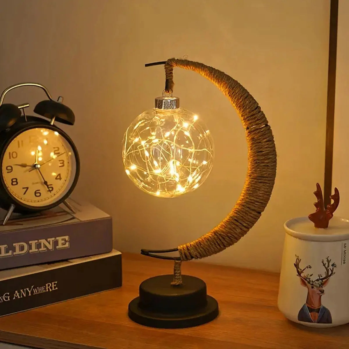 A cozy, modern table lamp designed in a crescent moon shape holds a glowing glass orb filled with warm fairy lights. The lamp sits on a wooden desk beside a vintage alarm clock, stacked books, and a decorative mug, creating a soft, relaxing atmosphere perfect for bedrooms or living spaces.